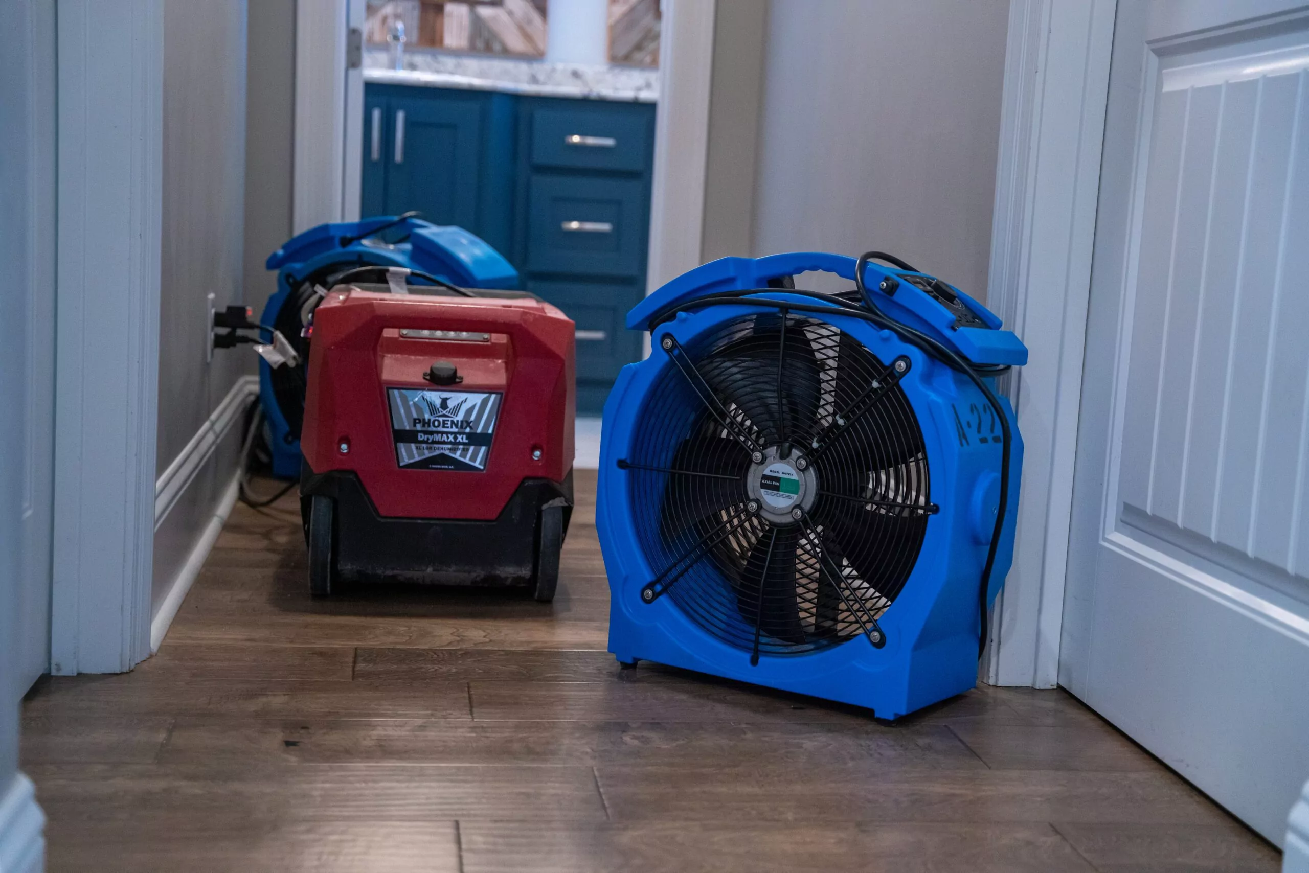 High-powered drying equipment, including a red Phoenix DryMAX XL dehumidifier and a blue air mover fan, set up in a hallway to dry hardwood floors after water damage.